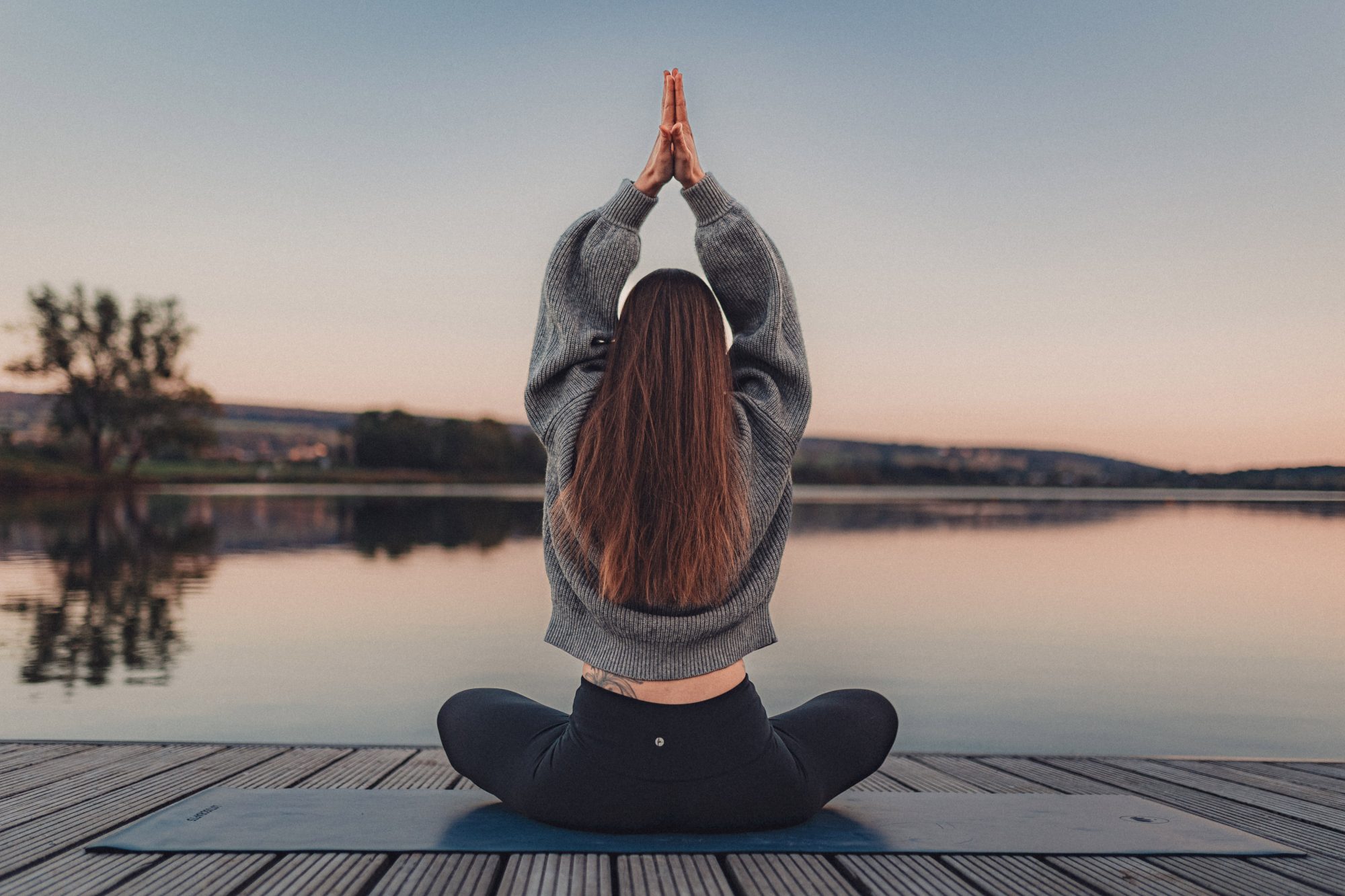 Helena Gnaase sitzt im Schneidersitz auf einem Steg vor einem See im Sonnenuntergang. Die Hände sind zum Anjali Mudra erhoben – eine Geste des Grußes und der Verbundenheit.
