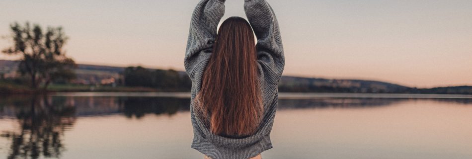 Helena Gnaase sitzt im Schneidersitz auf einem Steg vor einem See im Sonnenuntergang. Die Hände sind zum Anjali Mudra erhoben – eine Geste des Grußes und der Verbundenheit.
