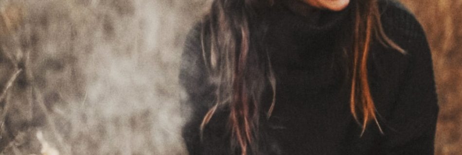 Yogalehrerin Helena Gnaase sitzt lächelnd mit einer weißen Tasse am Lagerfeuer. Umgeben von Natur, Herbststimmung und Rauch – ein Moment von Wärme, Achtsamkeit und Lebensfreude.