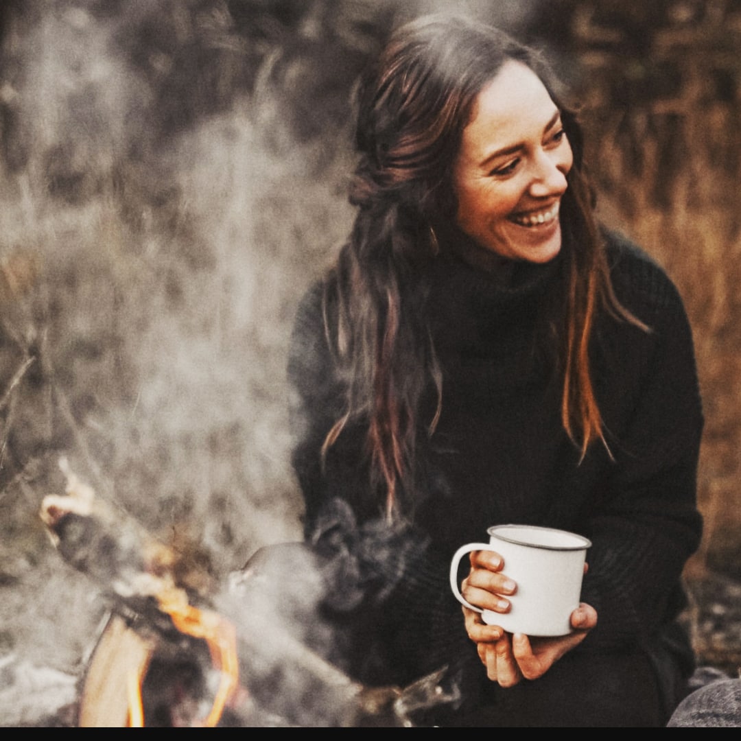 Yogalehrerin Helena Gnaase sitzt lächelnd mit einer weißen Tasse am Lagerfeuer. Umgeben von Natur, Herbststimmung und Rauch – ein Moment von Wärme, Achtsamkeit und Lebensfreude.