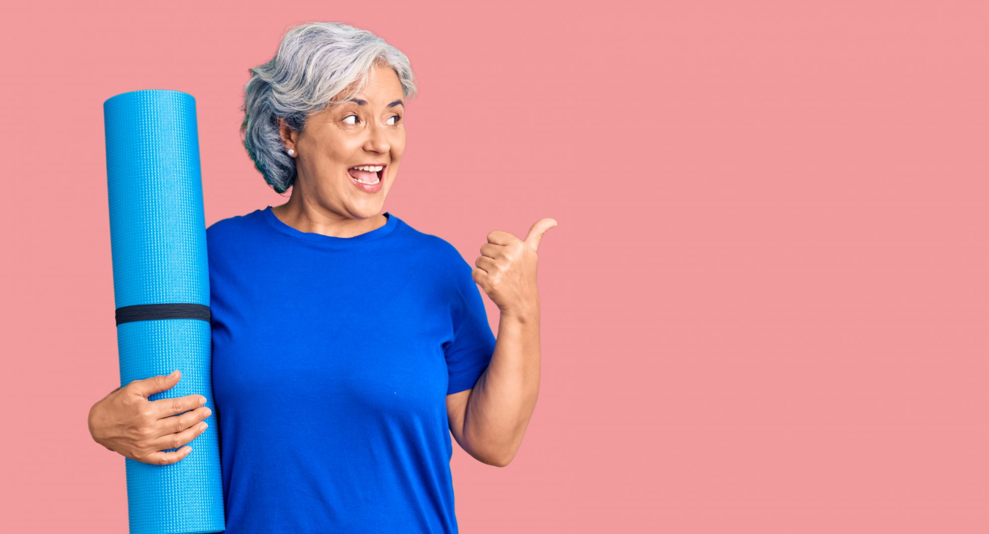 Eine lächelnde Frau mittleren Alters hält eine Yogamatte in der Hand und zeigt mit dem Daumen nach hinten – Symbol für Freude an Bewegung und barrierearmes Yoga.