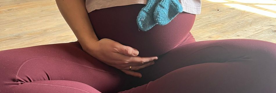 Schwangere Yogalehrerin Helena Gnaase sitzt im Schneidersitz und hält kleine Babysocken vor ihren Bauch – ruhige Yoga Praxis in Paderborn